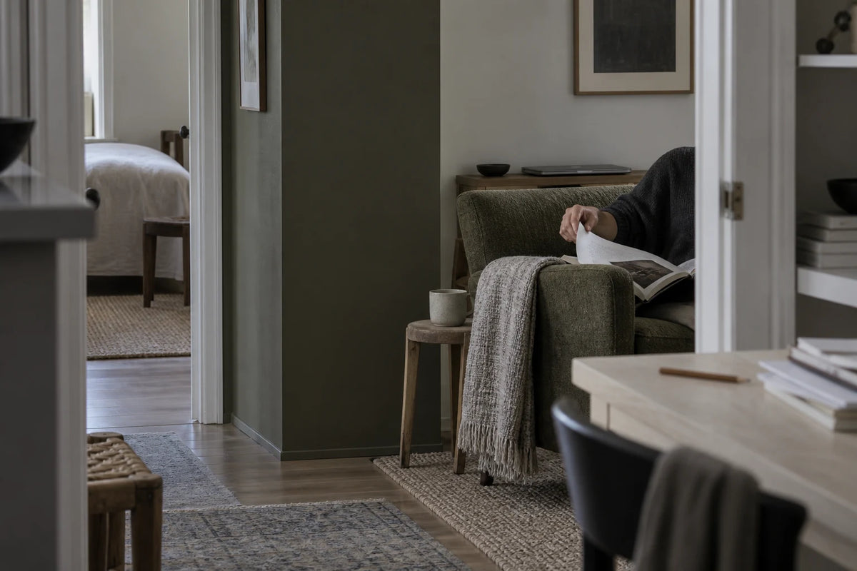 A cropped reader turns a page in an olive chair, with a closed laptop set aside behind it.