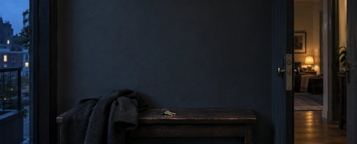 Keys and a wool coat set aside in a blue-grey apartment entry, between cool city light and a softly lit room.